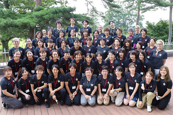 a large group of conference attendees posing in blue conference t-shirt with red nametag lanyards on an outdoor deck in fron tof various green trees