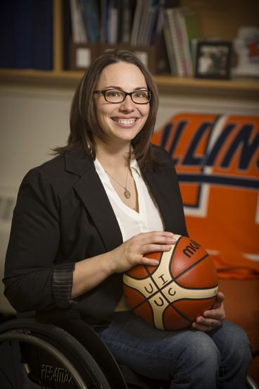 Stephanie Wheeler, smiling woman in glasses seated in a wheelchair in white top, blue blzer, jeans holding a basketball with the letters UIUC on it