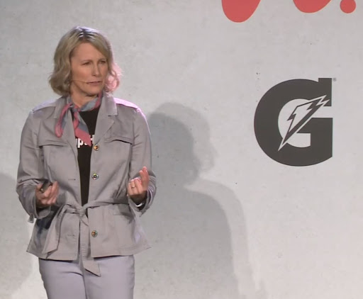 Nicole M. LaVoi, woman with shoulder-length blonde hair in gray suit, pink and blue neck scarf wearing a mic and gesturing to an audience with Gatorade logo visible